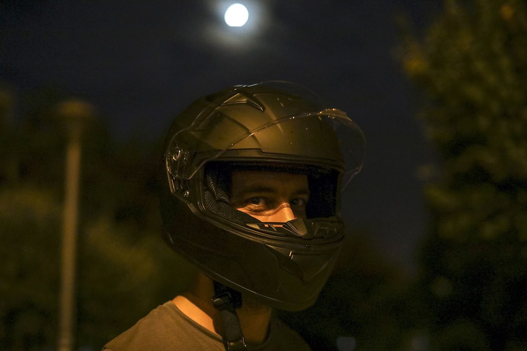 Henkilö musta moottoripyöräkypärä päässä katsoo kohti kameraa, taustalla öinen maisema kuutamoineen.